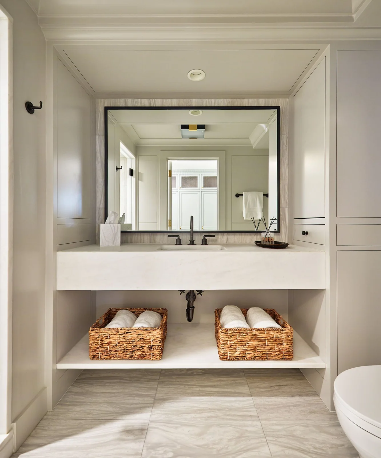 A broad pale basin counter stands over an open towel shelf and baskets beneath a black-framed rectangular mirror and pale paneled bathroom cupboards