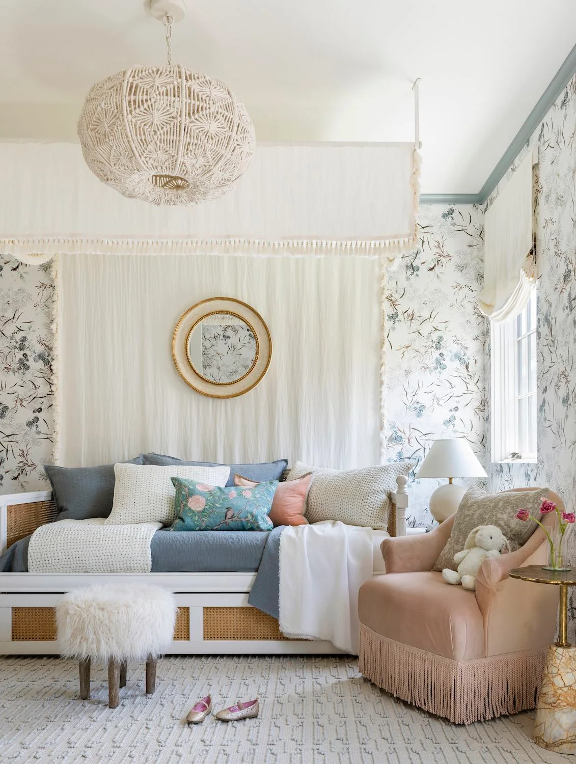 A pale blue daybed and pink fringed adult chair sit beneath botanical wallpaper, a woven pendant and hanging fabric beside a patterned nursery rug