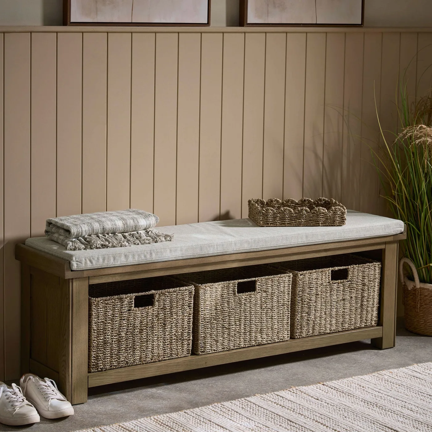 Wood storage bench with pale padded top and three woven baskets beneath, set against beige vertical paneling
