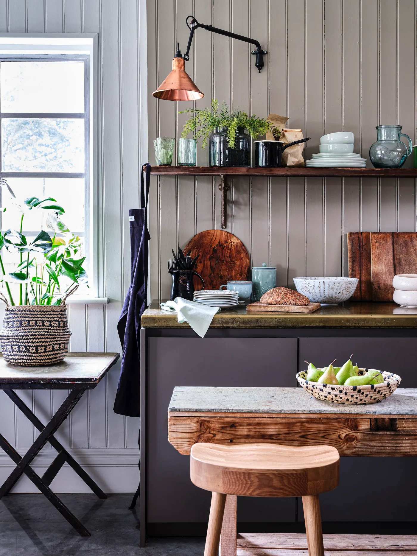 Cottage kitchen corner with gray vertical paneling, darker lower cabinets, wooden shelf, wooden table edge and warm metallic wall lamp beside a window.