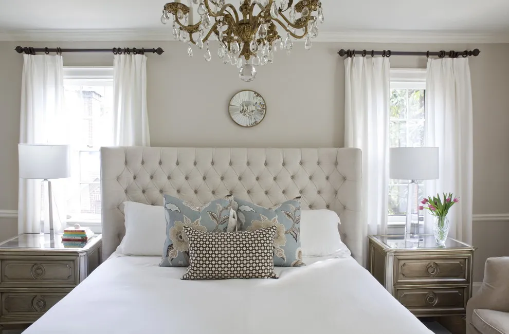 A tall tufted beige headboard sits beneath pale walls and a decorative light