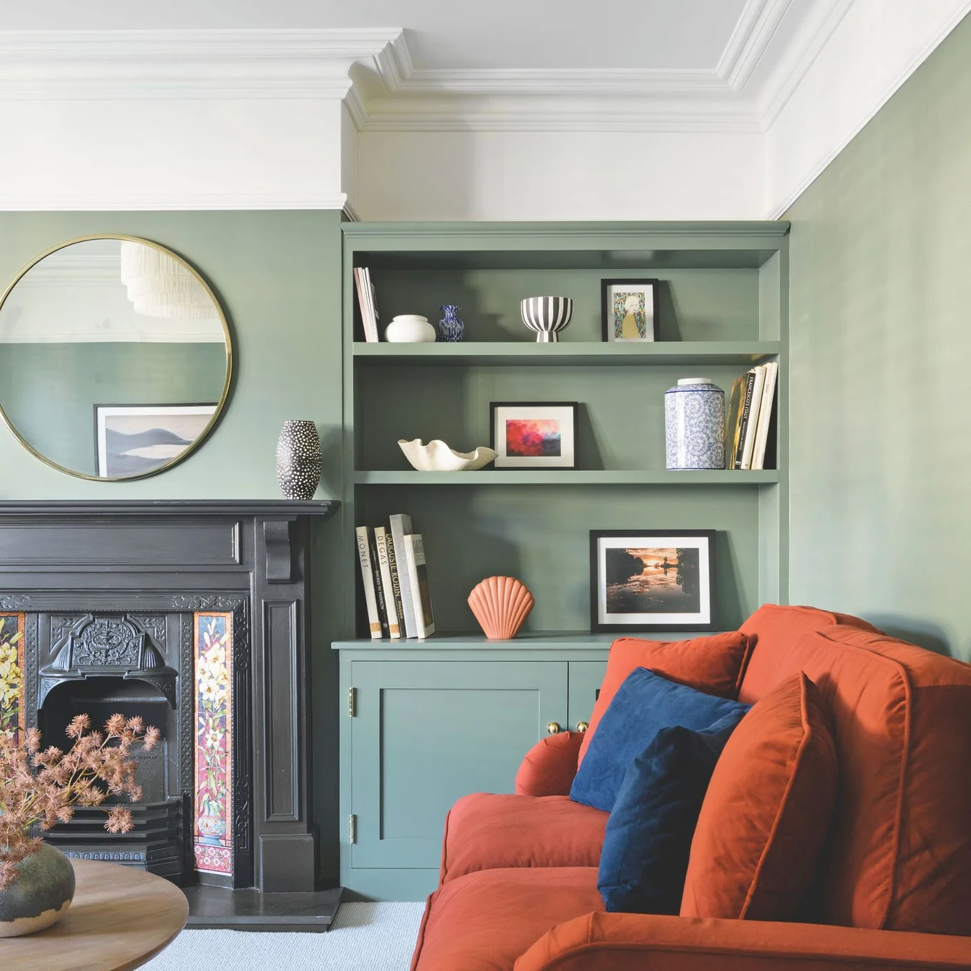 Sage green walls and open shelving frame an orange sofa beside a dark fireplace and round mirror