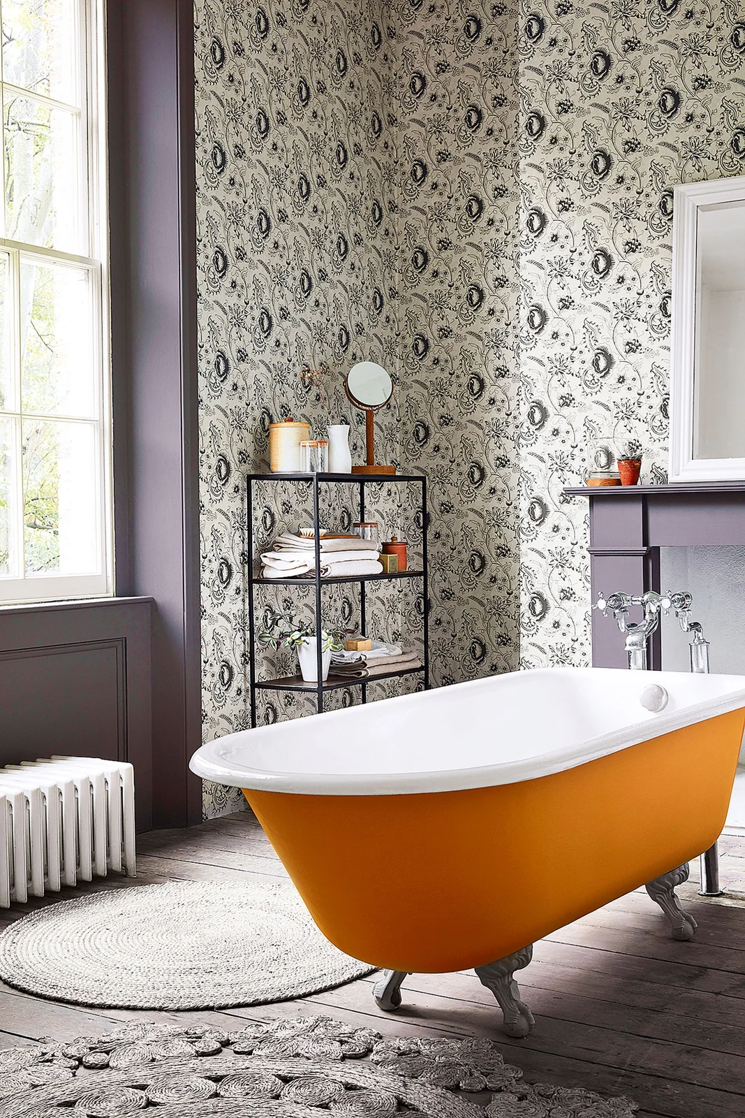 An orange claw-foot bath sits before densely patterned wallpaper and dark plum trim beside open shelves and a pale round rug