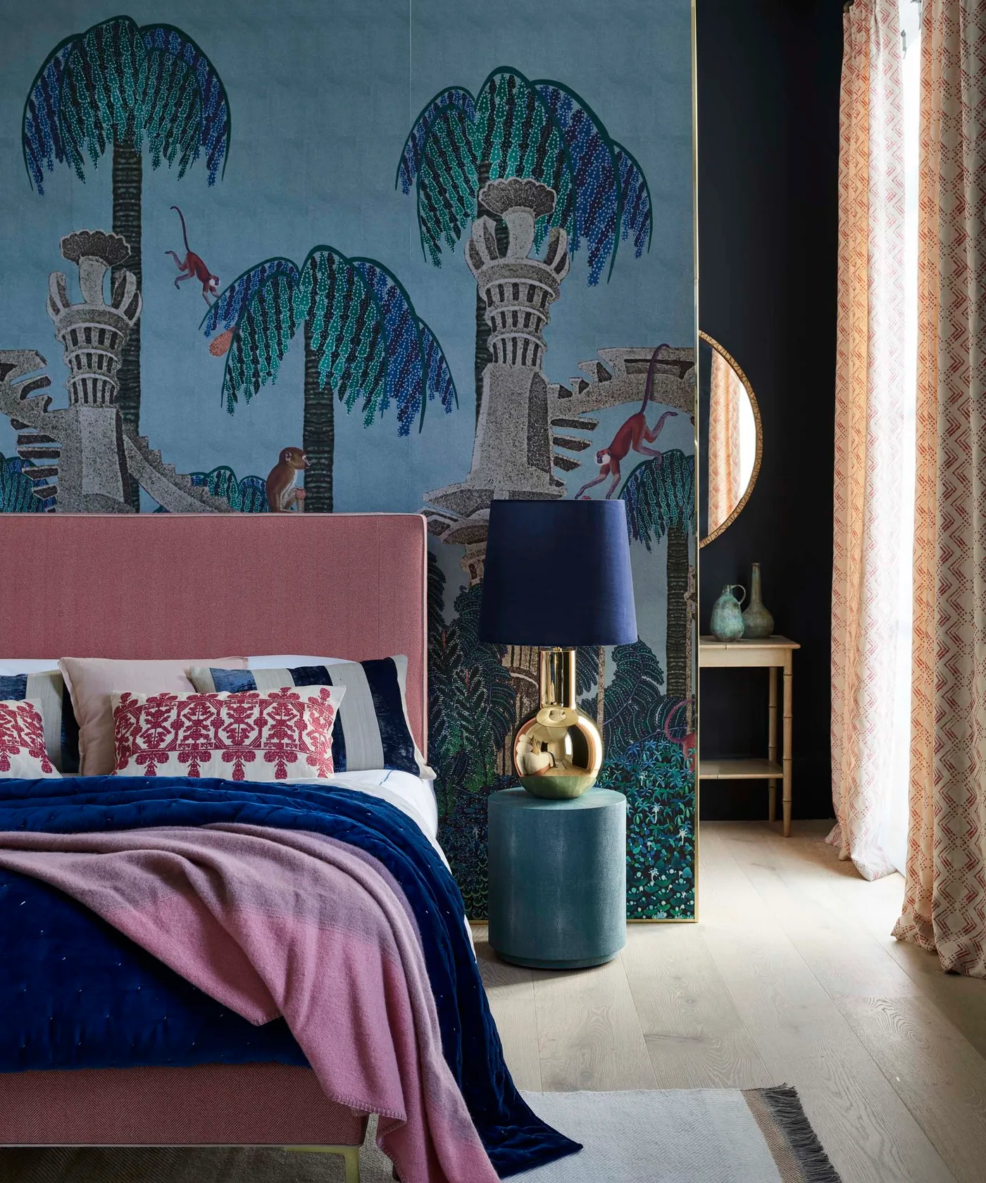 Blue-gray leafy bedroom wallpaper with a pink upholstered bed, dark blue bedcover, navy lampshade, and turquoise bedside table.
