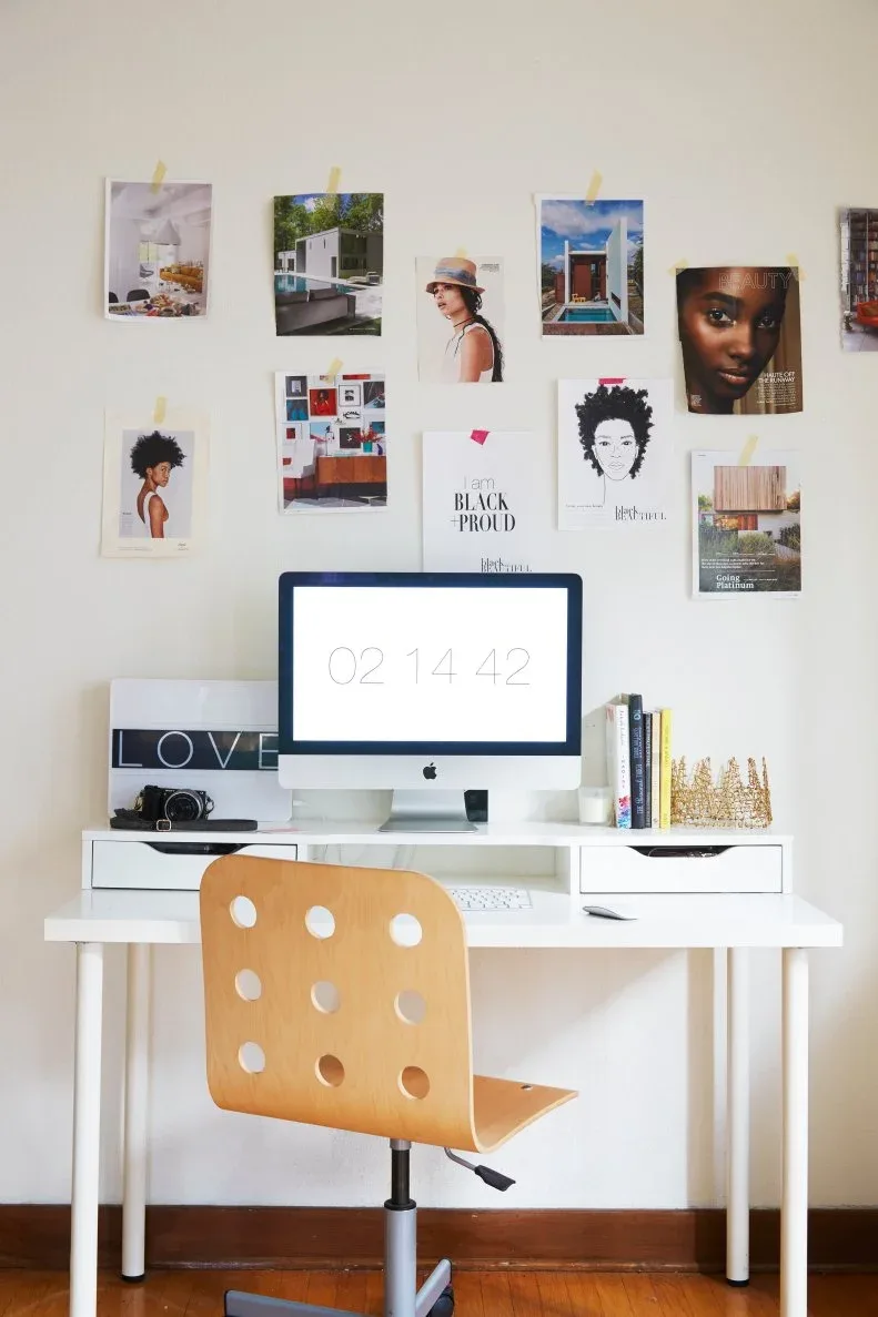 Home workspace with white drawer desk, computer, warm wooden chair, small books and decorative objects, and an informal wall display of photographs and illustrations.