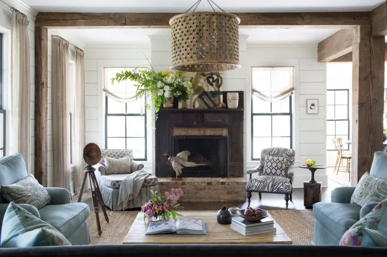 Country living room with pale blue sofas, patterned chairs, dark fireplace, broad timber framing and large patterned pendant