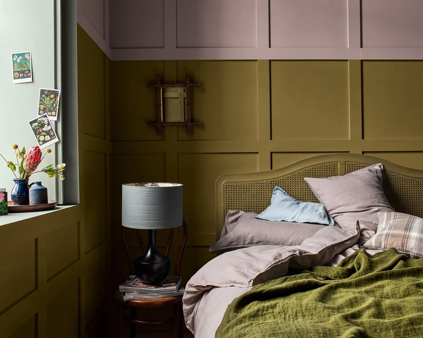 Bedroom with olive lower paneling, muted pink upper paneling, matching woven headboard, pale pink bedding, green throw, and small bedside lamp.