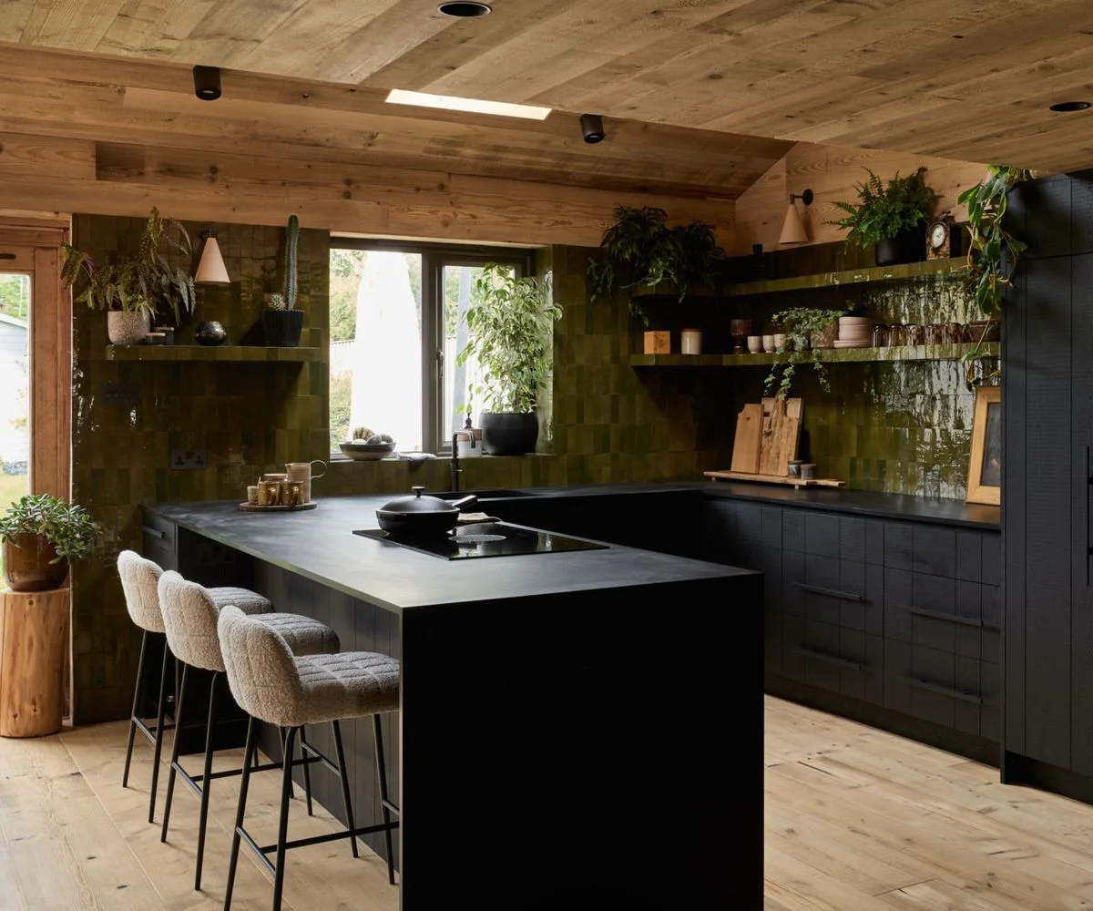 A black island and lower cabinets sit beneath a wooden ceiling beside green tiled walls, open shelves, plants and cushioned stools