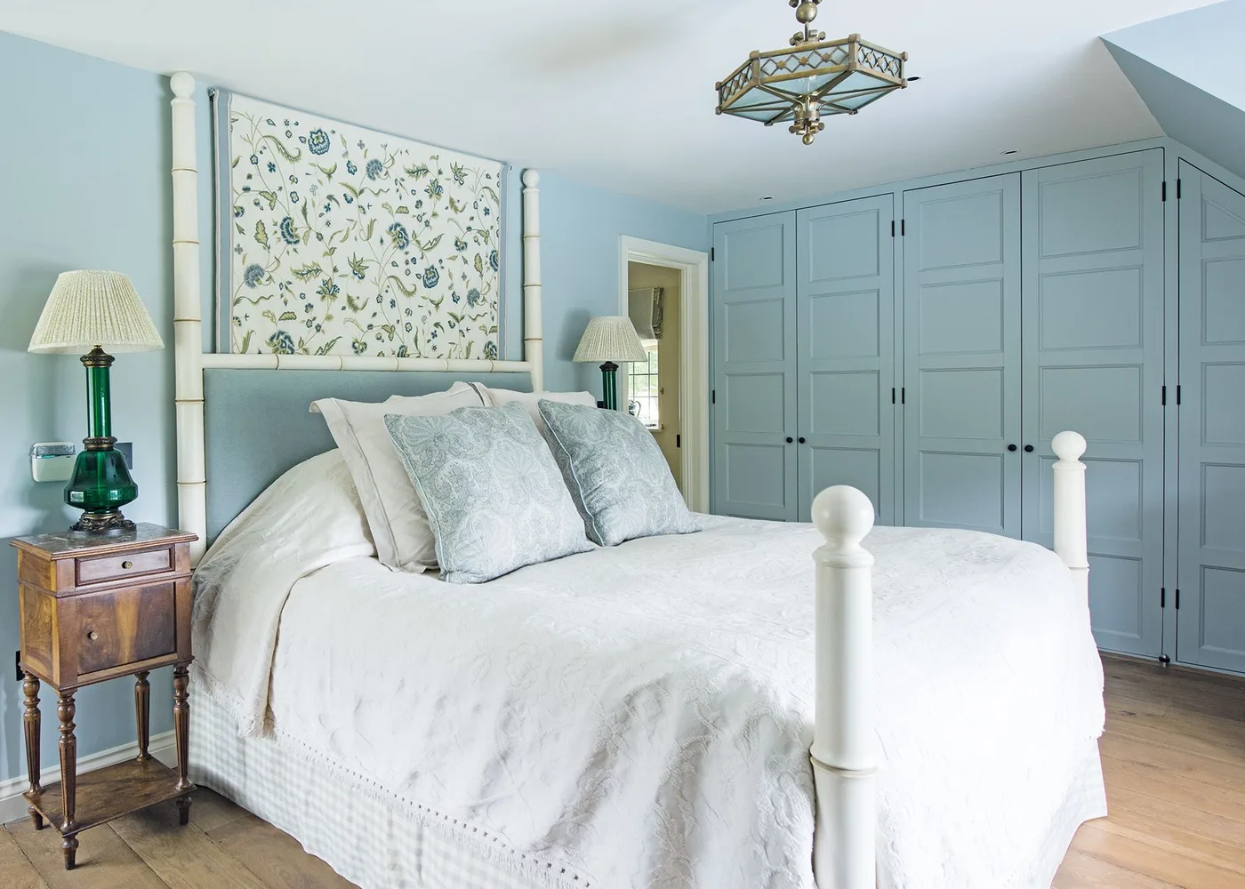 Pale blue bedroom with fitted paneled cupboards, a white bed frame, floral fabric behind the headboard and a small wooden bedside cabinet.