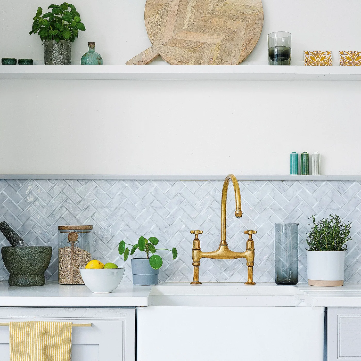 Pale rectangular tiles form a herringbone backsplash behind a white sink and gold-toned tap beneath a long white shelf