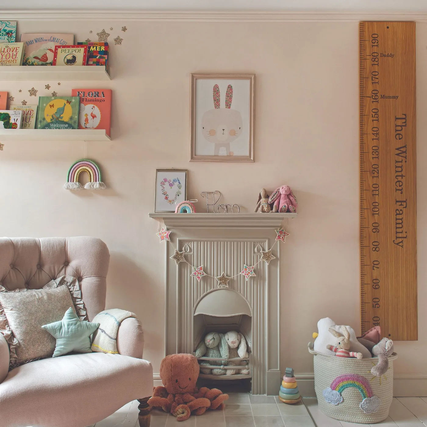 Pale pink adult chair beside cream fireplace with small framed animal pictures, books and tall wooden measuring piece.