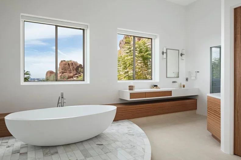 A white oval bathtub sits on a curved tiled platform beside white walls, two rectangular windows and a long floating wood-fronted vanity