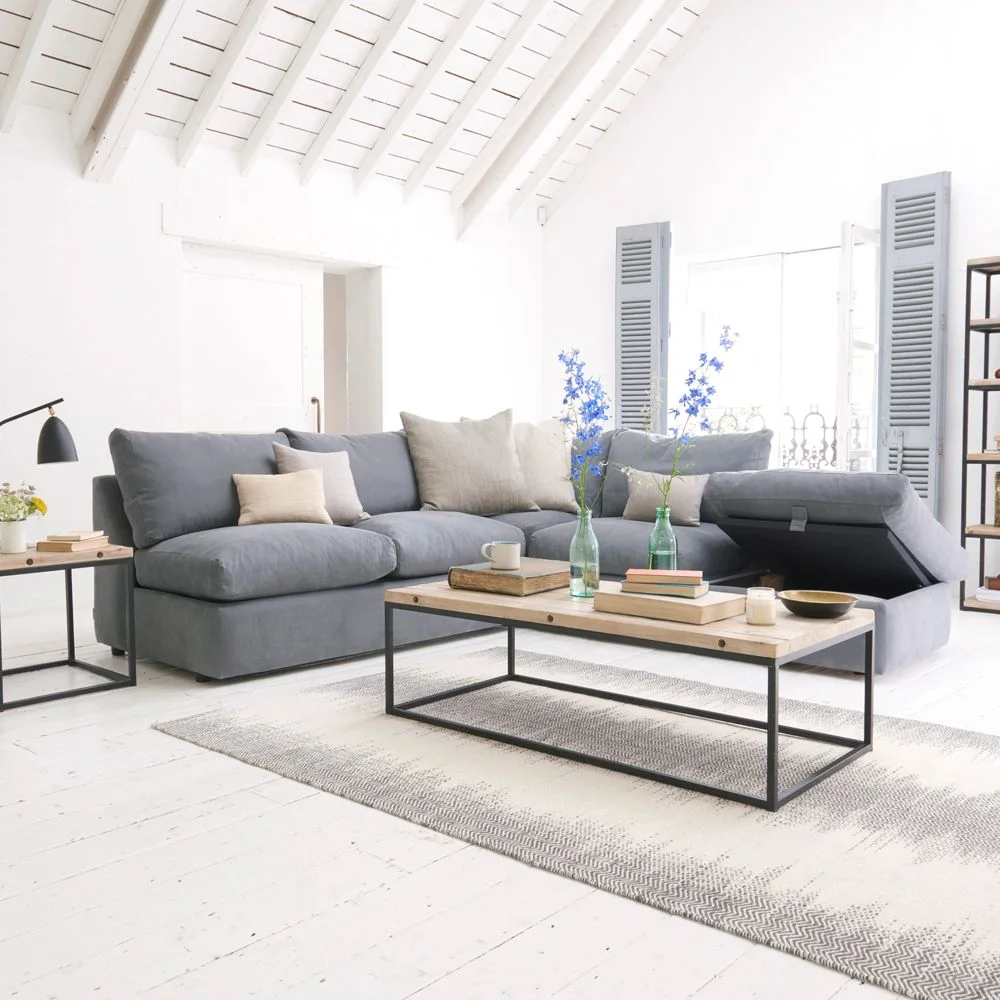 A gray corner sofa has its chaise storage lid open in a white-roofed room beside a rectangular wooden-topped coffee table