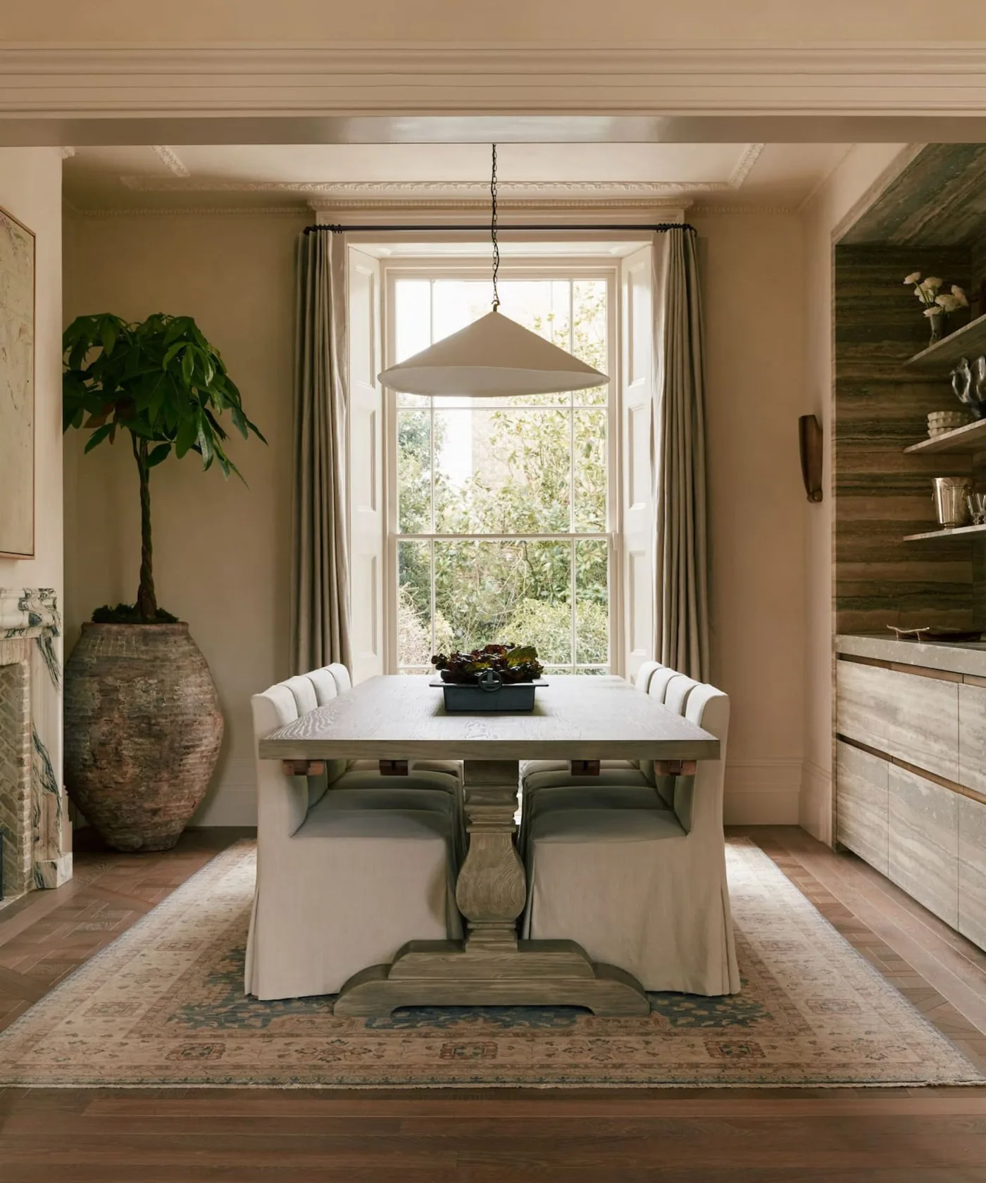 Cottage dining room with warm pale walls, wooden pedestal table, pale skirted chairs, recessed wooden storage and a plant in a large textured container.