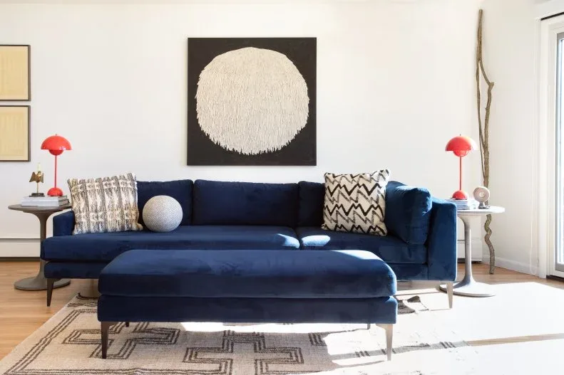 Living room with dark blue sofa and matching ottoman, red dome-shaped lamps on pale side tables, patterned cushions and a large circular abstract artwork.