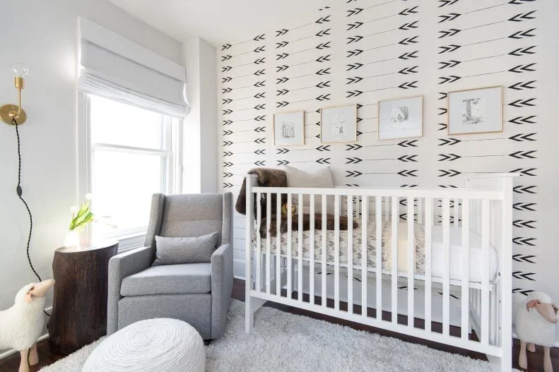 A white nursery crib stands beside a gray adult chair and white rounded footrest below repeated dark arrow shapes on a pale wall