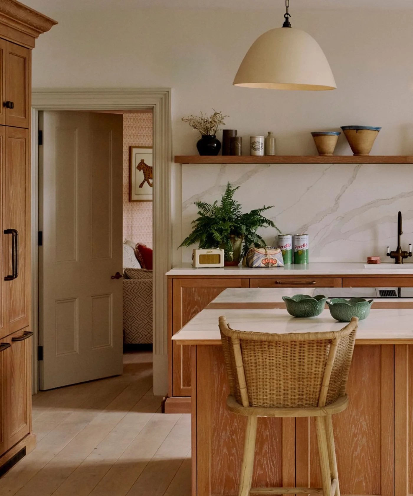 Wooden kitchen cabinets with a pale veined backsplash, black faucet and long wooden shelf holding bowls above a white counter.