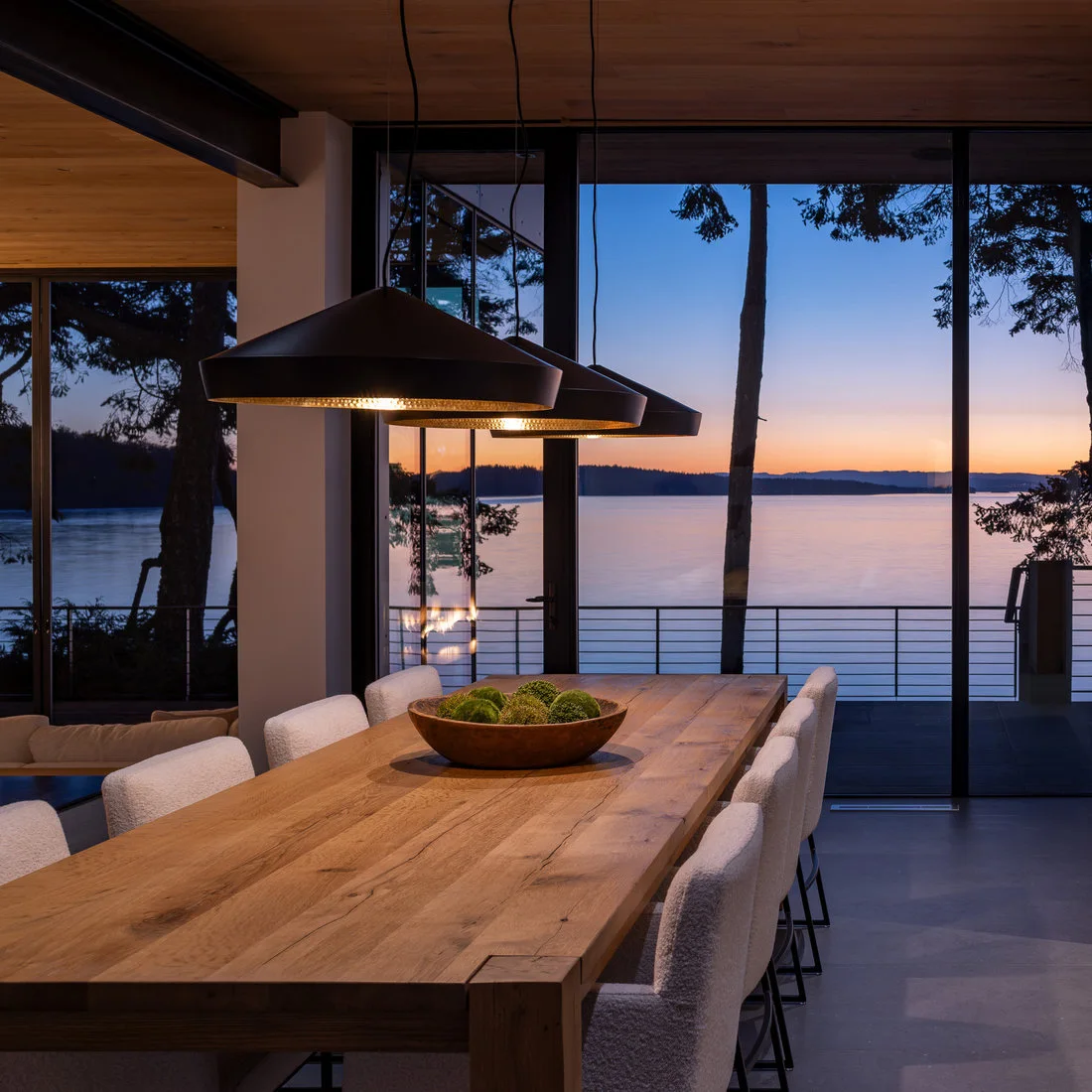 A long wood table and pale upholstered chairs sit under three dark angular pendants beside a wood ceiling and tall black-framed windows overlooking water