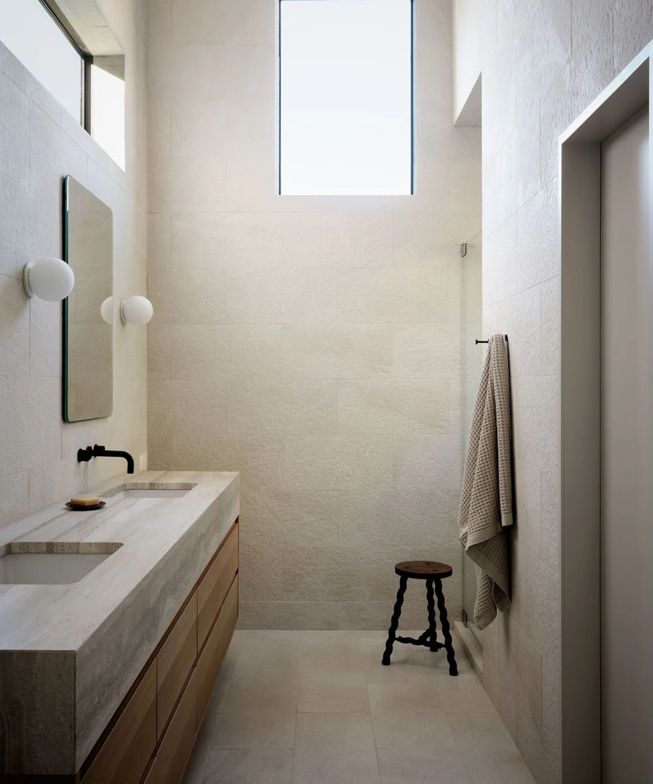 A long wood-fronted double vanity with a pale veined counter stands beneath a rectangular mirror and white globe lights beside textured pale walls and a dark stool