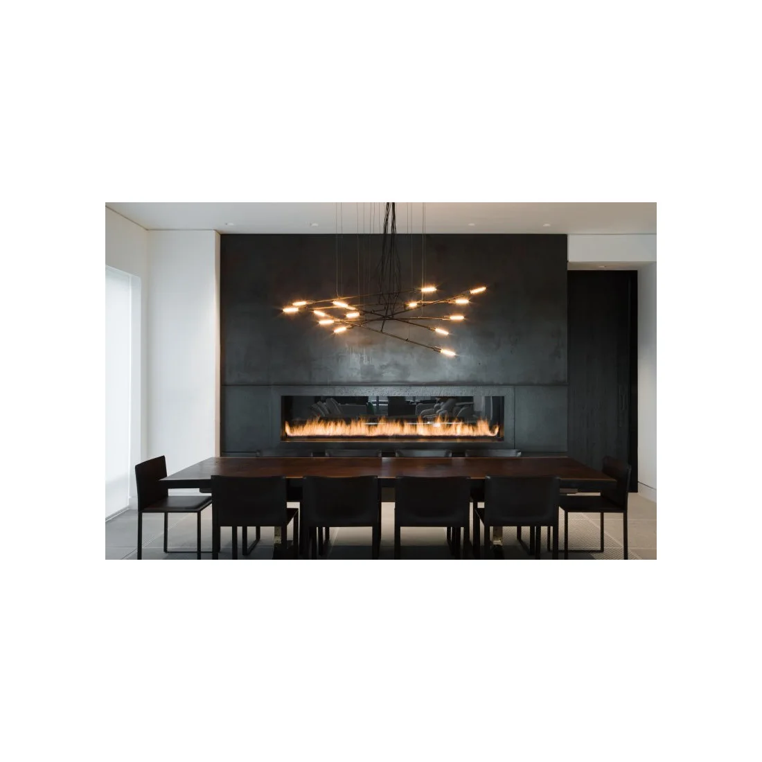 A dark rectangular dining table and chairs sit beneath a branching light beside a charcoal wall with a long horizontal fireplace