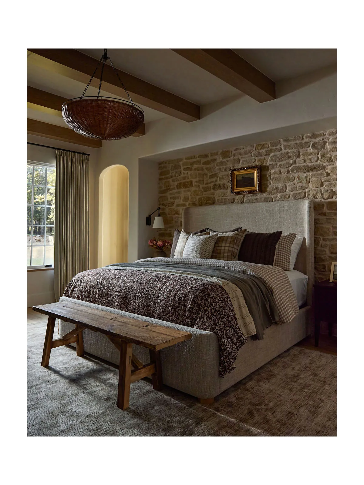 A timber bench stands at the foot of a pale upholstered bed against a stone wall beneath exposed beams.