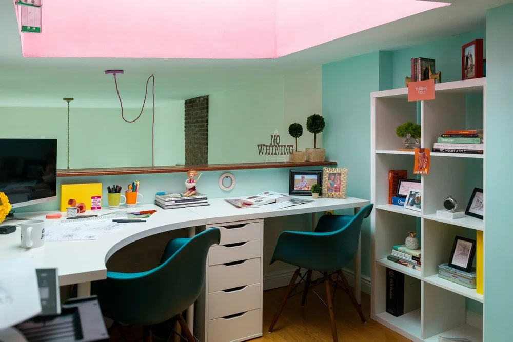 A long pale work surface with two teal chairs sits beneath mint green walls beside open cubby storage and a pink ceiling area