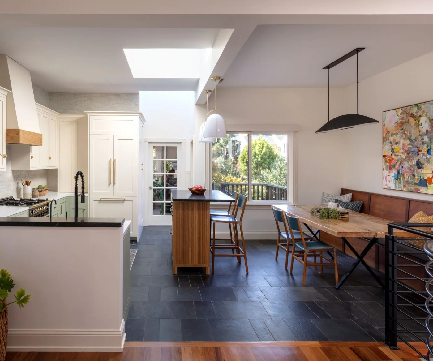 A wooden dining table and wall bench stand alongside a wooden island in a long pale kitchen with dark floor tiles
