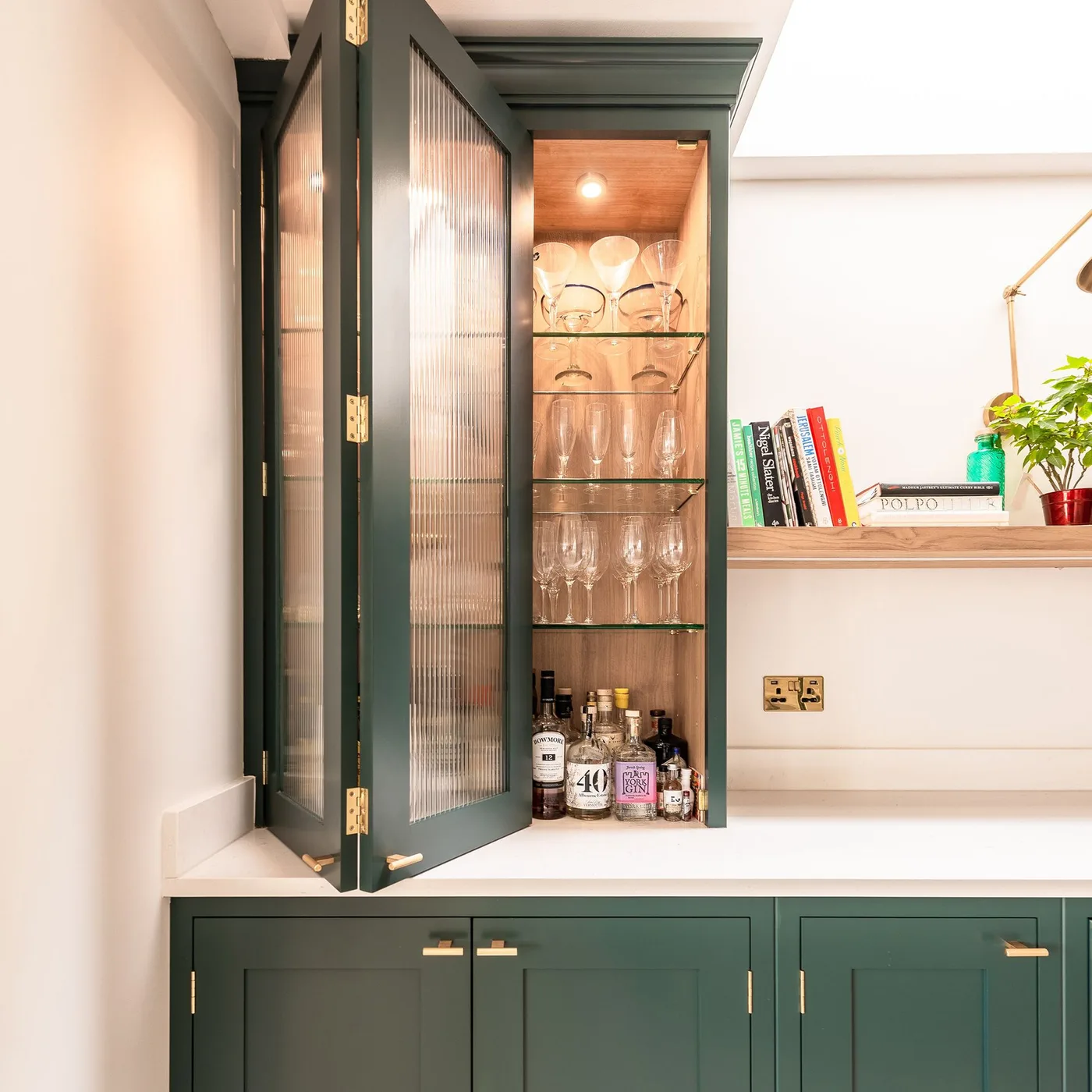 Green framed kitchen cupboard with textured glass doors, lit glass shelves and bottles below a glassware display.