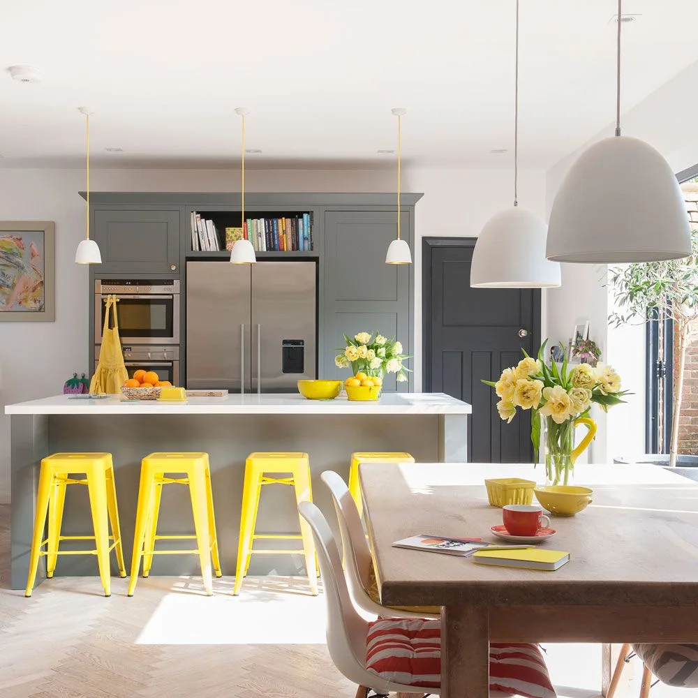 Yellow stools line a pale island beside a wooden dining table, white pendant shades and gray kitchen cabinets