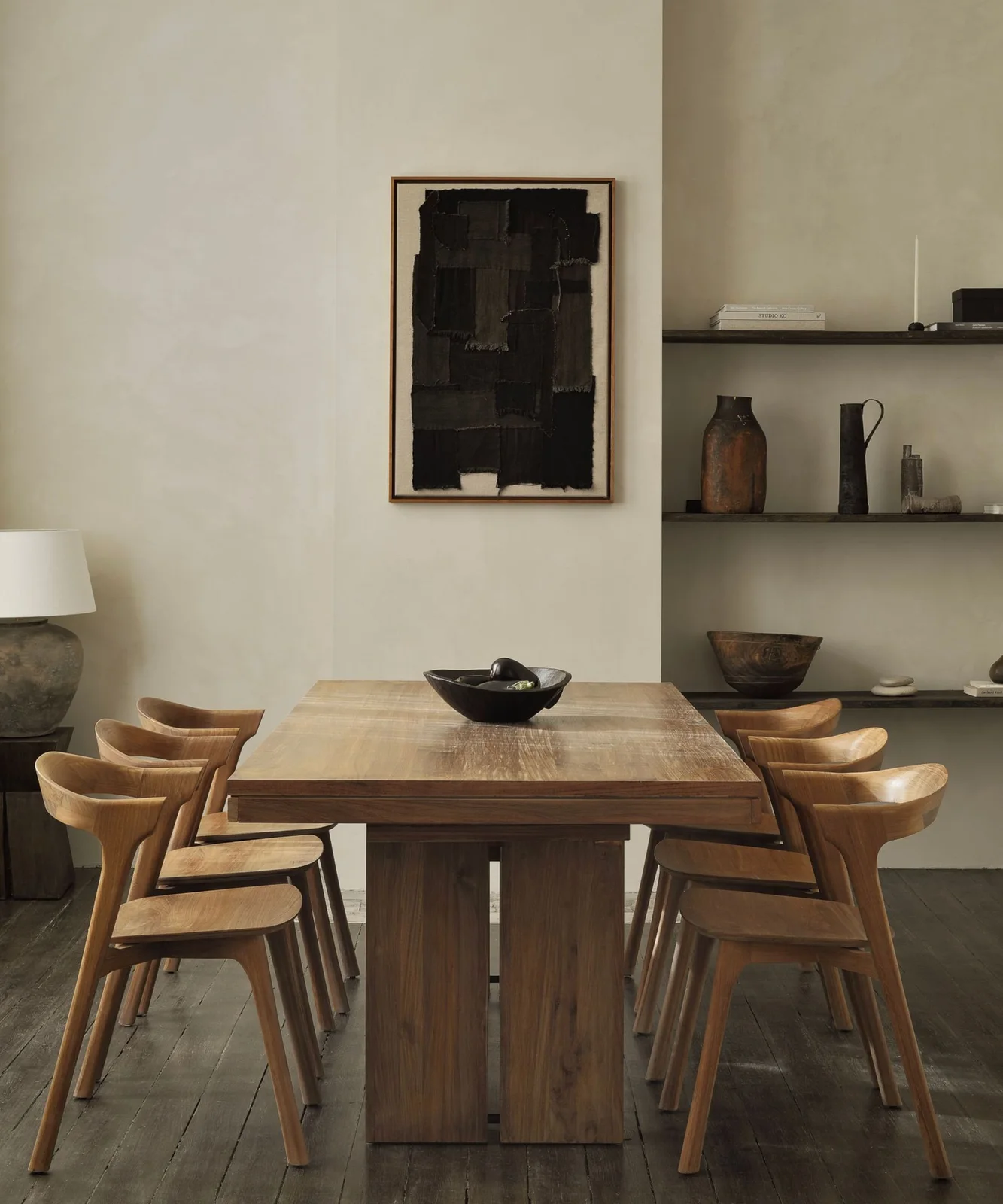 Sculpted wooden armchairs surround a broad wooden dining table beside dark open shelves and a large black artwork
