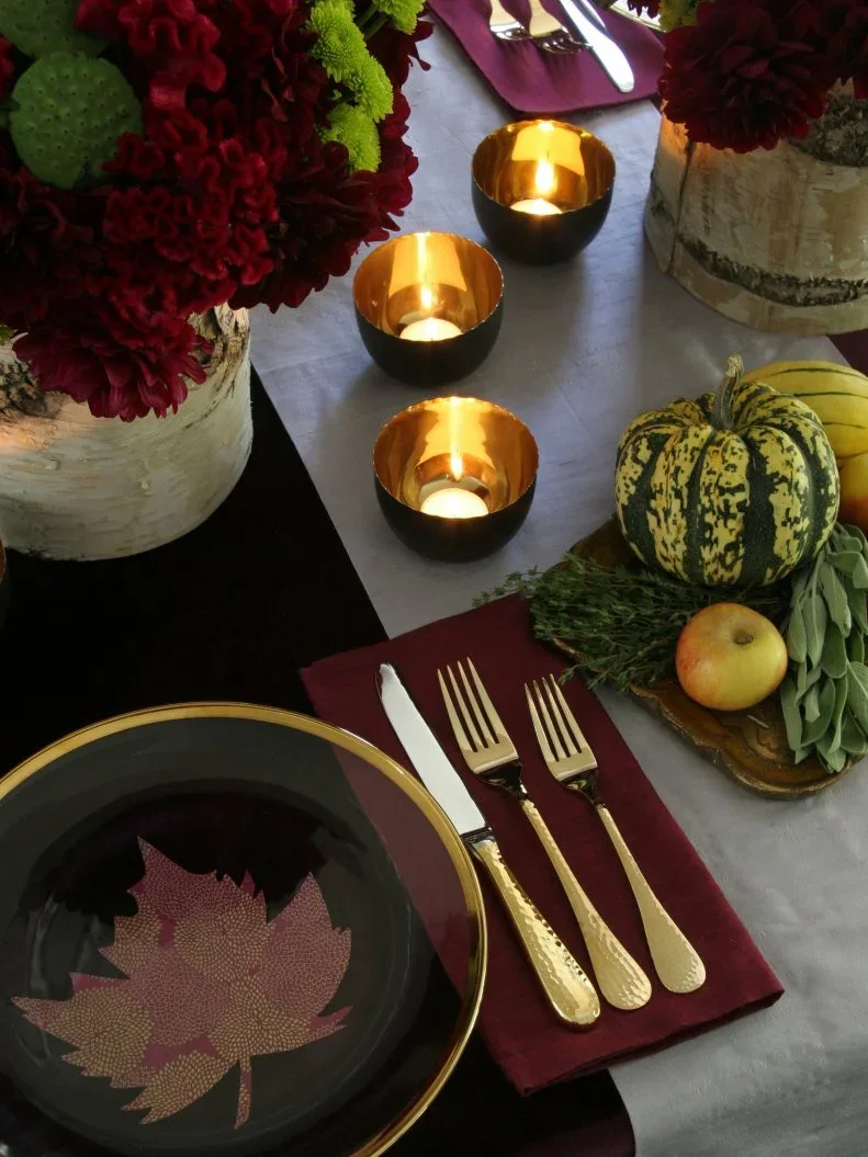 A dark plate with a warm metallic rim holds a decorative leaf shape beside gold-colored cutlery on a plum napkin