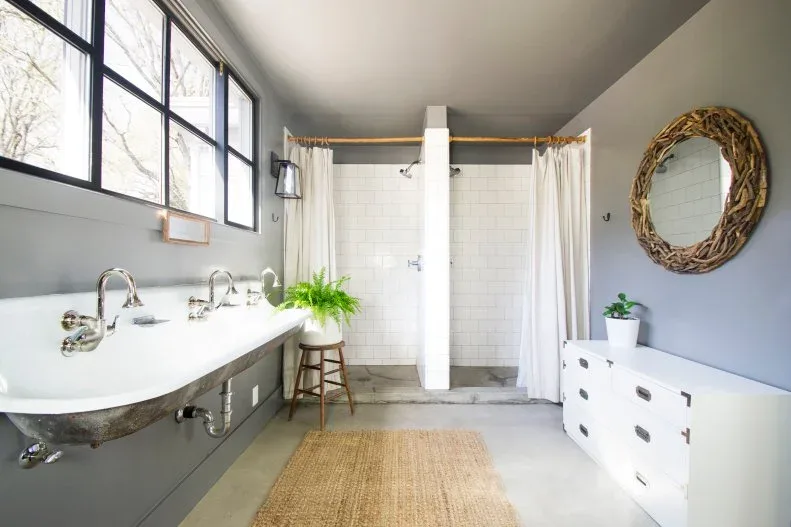 Gray paneled bathroom with long white basin and three taps, high black-framed windows, two curtained shower bays and woven mirror