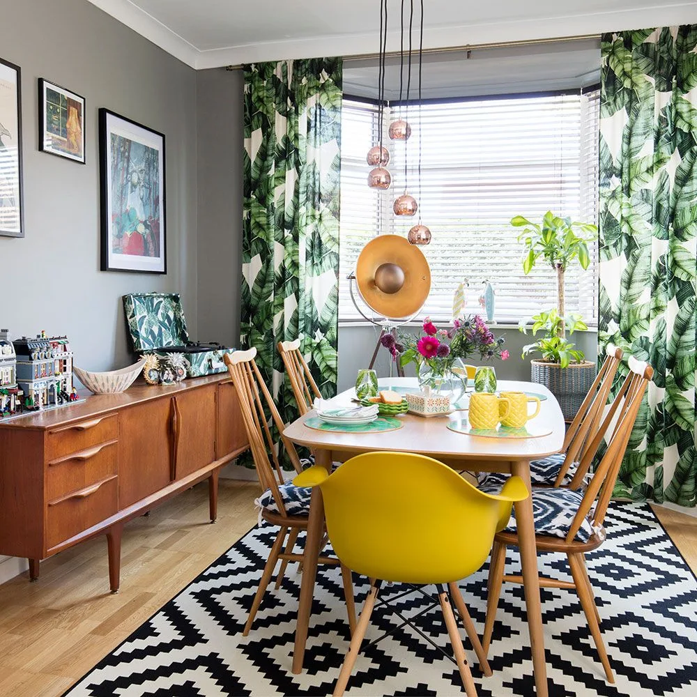 Green leaf-print curtains framing a window with a pale horizontal blind, beside gray walls, a wooden table, yellow chair, and black-and-white geometric rug.