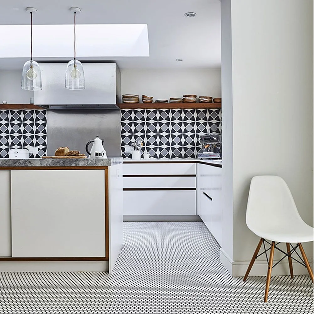 A white kitchen has black-and-white patterned wall tiles and a small checked floor below pale cupboards beside a white chair and clear pendant lights