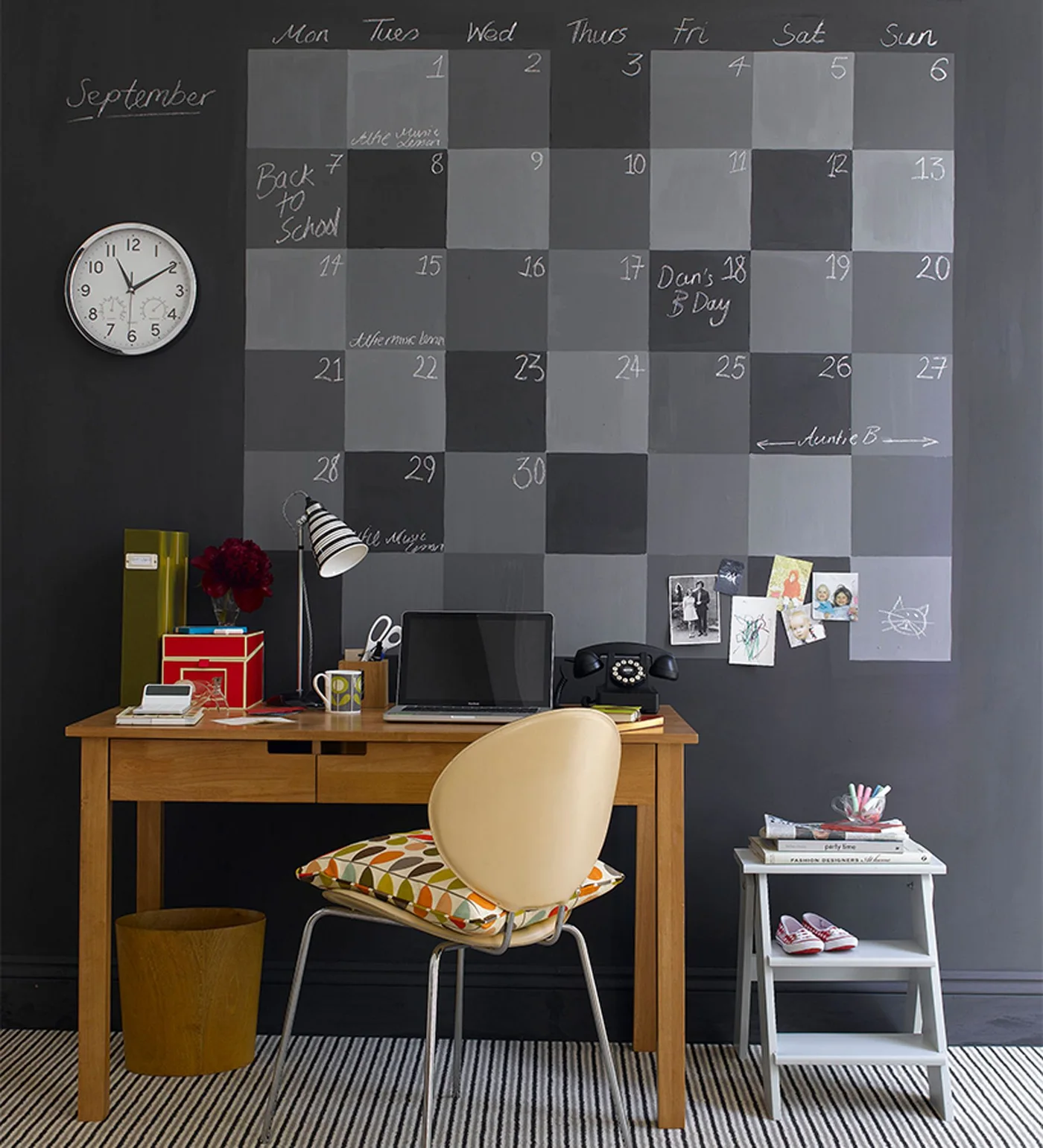 A wooden desk and pale chair stand beneath a large gray chalkboard calendar beside a white clock and a small white side shelf