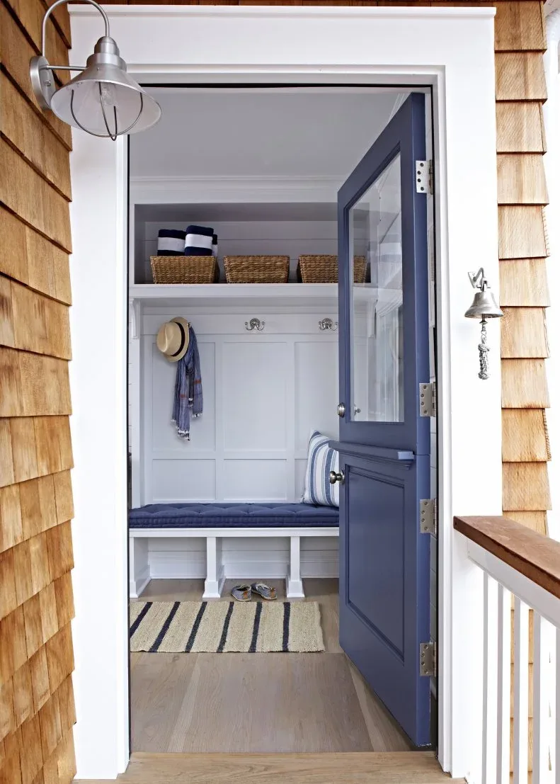 Open navy entrance door showing white paneled mudroom with blue cushioned bench, upper baskets and striped mat.