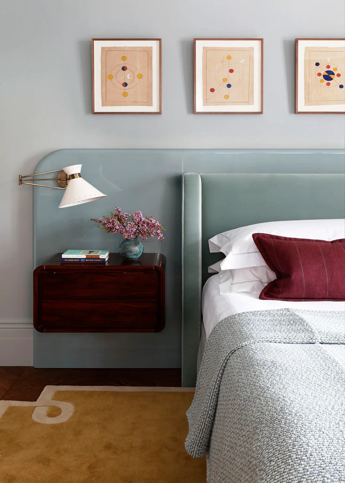 A floating dark wood bedside cabinet sits against a pale blue headboard beside a wall light, framed prints and burgundy cushion