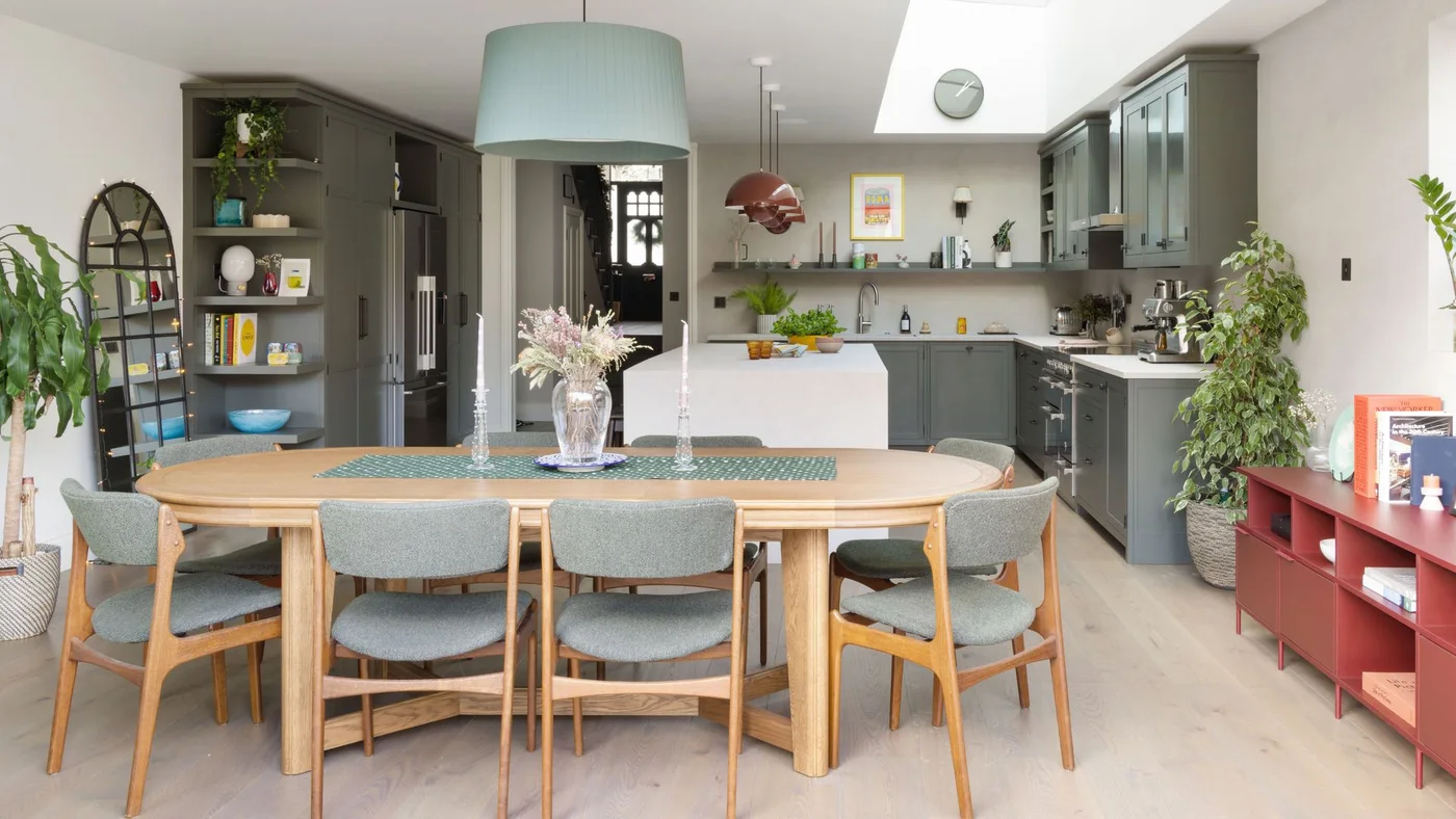 An oval wooden table with gray upholstered chairs stands in front of a gray kitchen and pale island beneath a large pendant