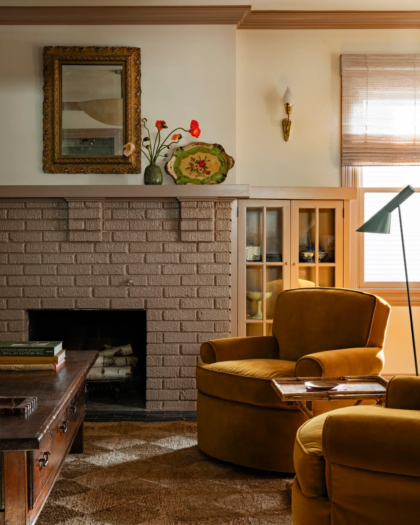 Warm wood glass-front cabinet beside painted brick fireplace and ochre armchair