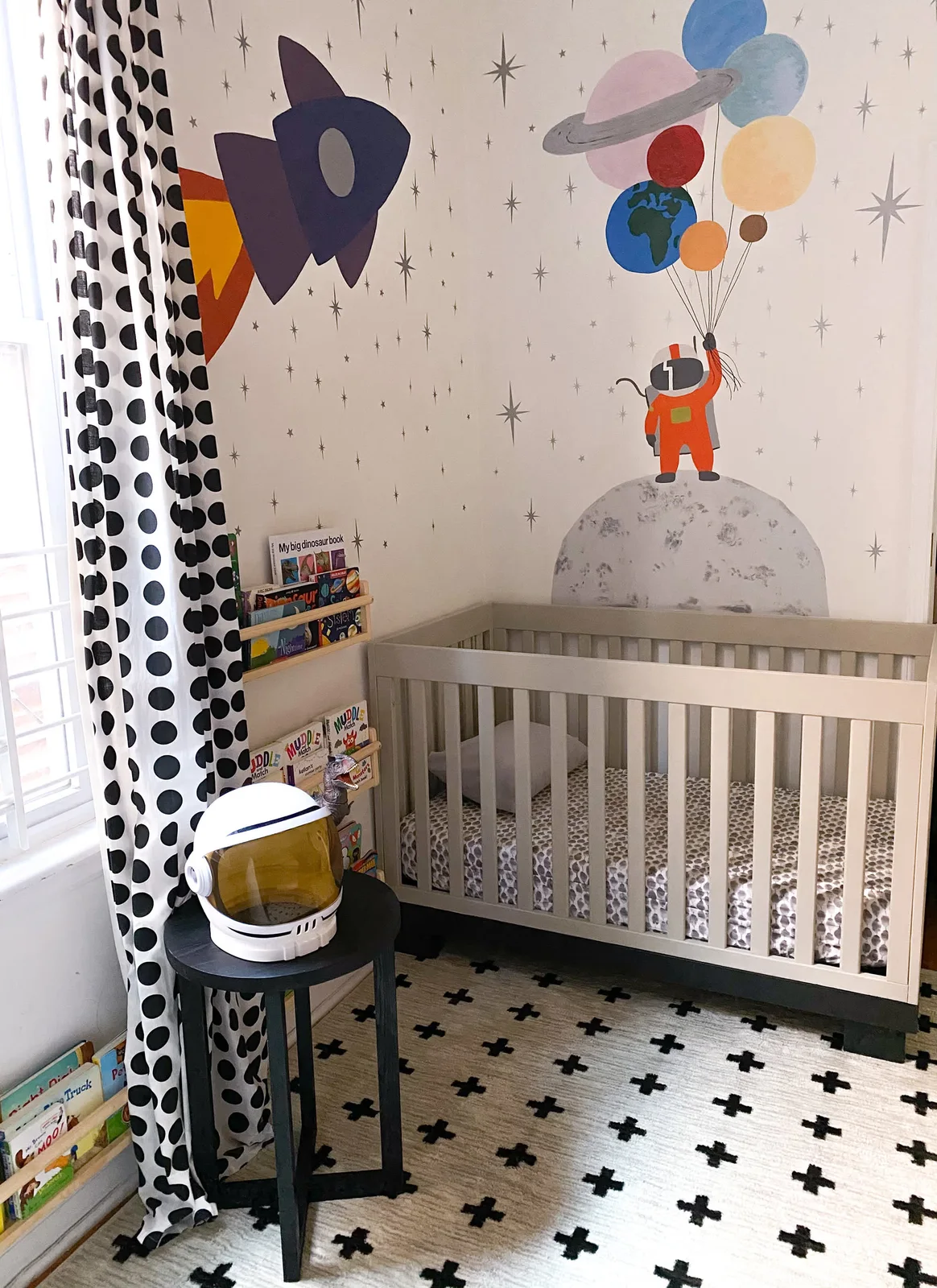 A pale nursery crib stands beside black-and-white graphic curtains and a pale rug with black crosses below a colorful space-themed wall mural