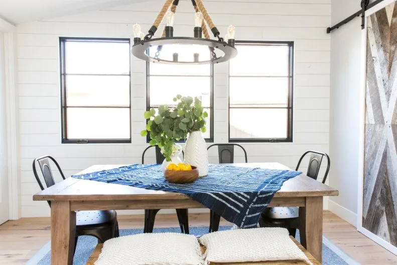 Blue patterned cloth laid diagonally under a white vase of foliage and a bowl of yellow fruit on a wood table