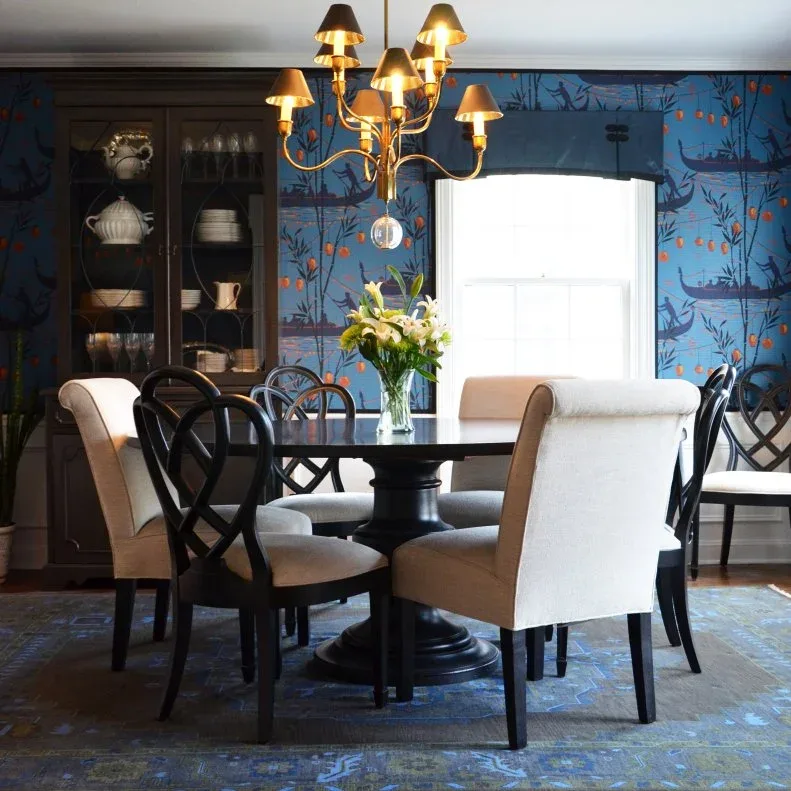 Gold-toned chandelier with small dark shades and a clear hanging sphere above a dark round table, in a blue wallpapered dining room with pale end chairs.