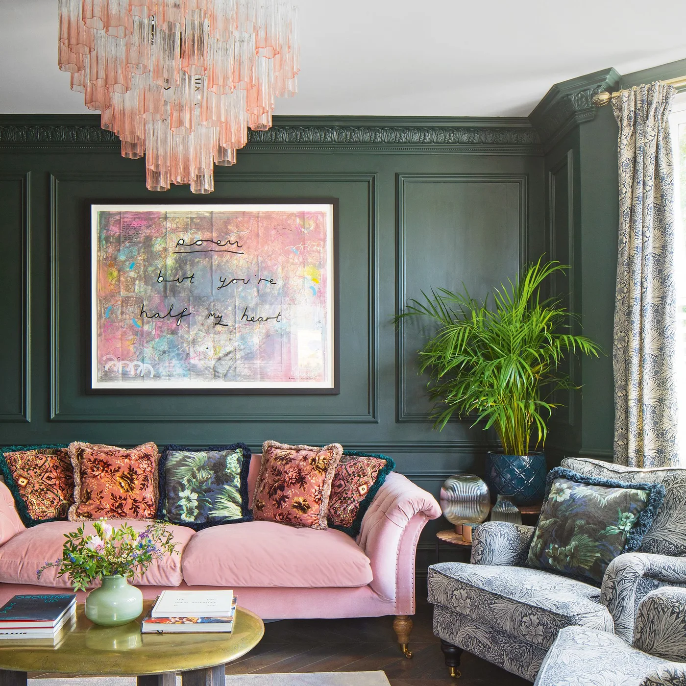 A pale pink sofa and pink glass chandelier sit against dark green paneled walls beside a patterned chair