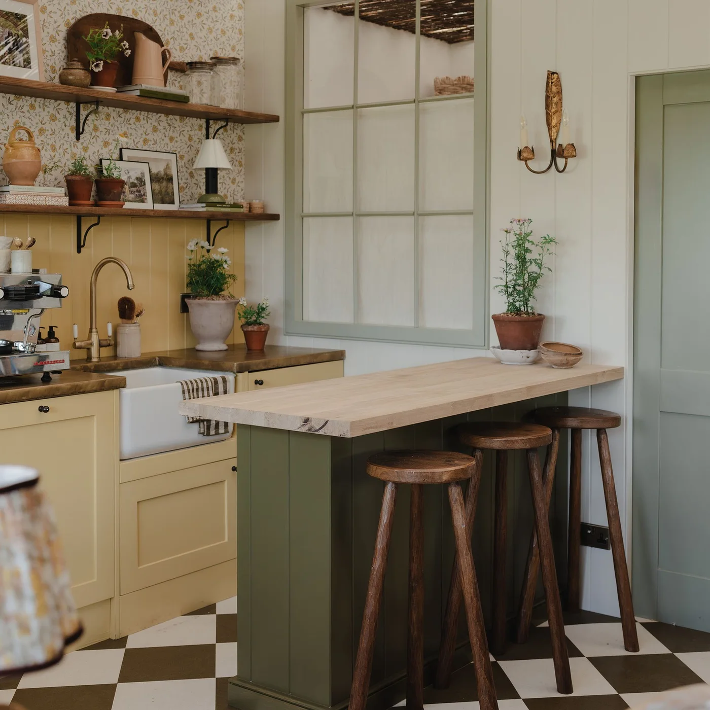 A deep green peninsula with wooden stools sits beside pale yellow lower cabinets, wooden shelves and a pale sink