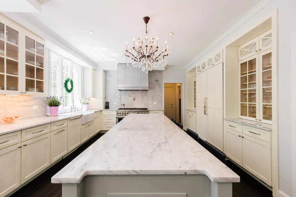 Pale paneled cabinets with glass uppers line a long kitchen beside a light island