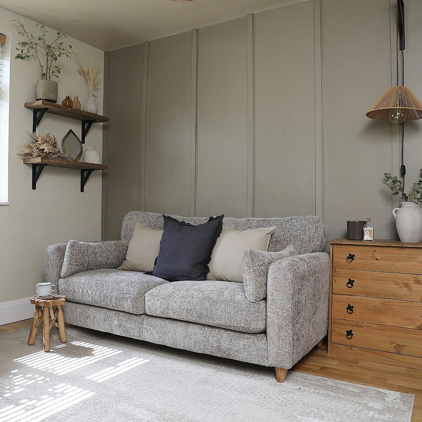 A gray sofa sits beside wooden drawers beneath a paneled gray-beige wall, wooden shelves and a woven lamp shade