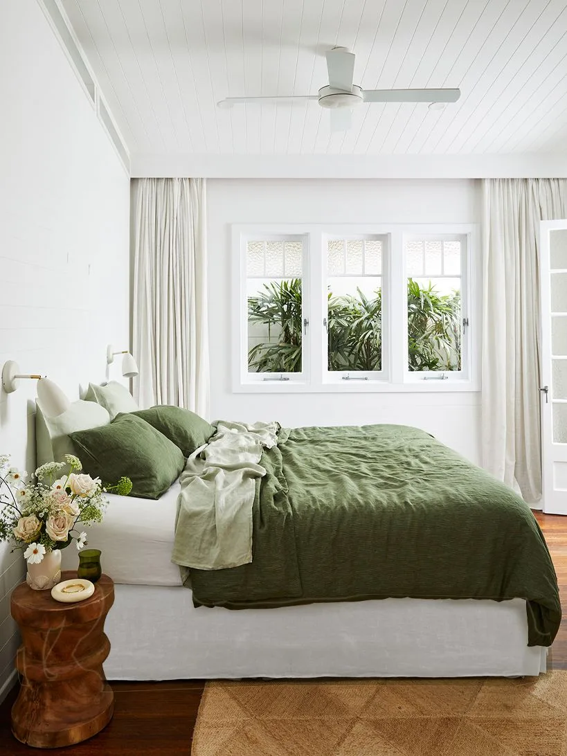 Bright white bedroom with olive bedding, a spiral-shaped wood bedside table, woven rug, small-paned windows and white curtains