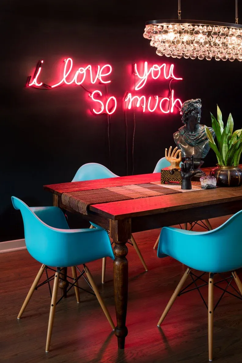 Pink-red illuminated script on a dark dining-room wall, with a wooden table, bright blue chairs, suspended glass globes, a dark bust, and a plant.