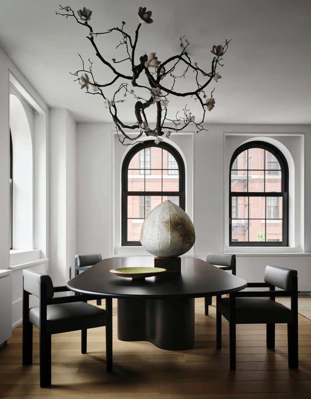 Black dining chairs and a dark pedestal table stand beneath a branching light beside pale walls and black-framed arched windows