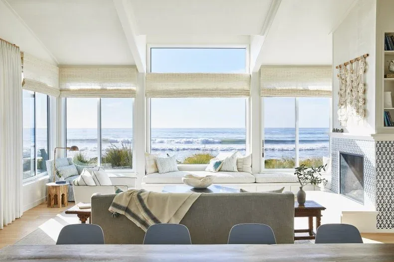 Living room with pale low sectional by broad ocean-facing windows, gray sofa back, wooden tables, pale woven shades and patterned fireplace surround.